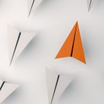 one orange paper airplane going in the opposite direction of all the other white airplanes